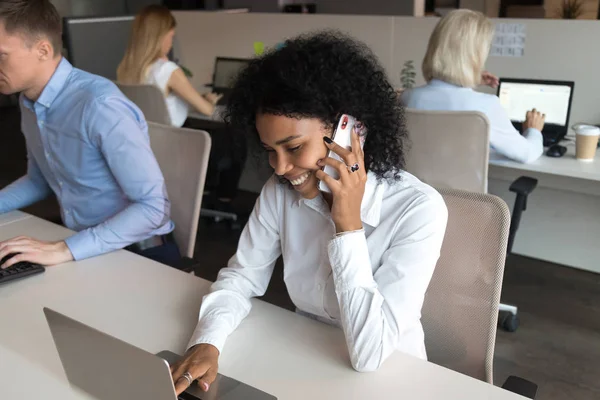 Sonriente charla de empleados negros en el teléfono inteligente que trabaja en el ordenador portátil — Foto de Stock