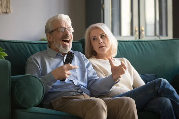 Annoyed elder woman looking at cheering senior husband, watching TV. — Stock Photo, Image