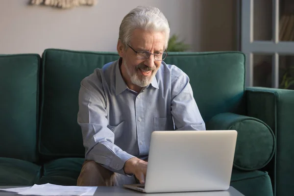 Sorrindo homem idoso usando laptop navegando na internet em casa — Fotografia de Stock