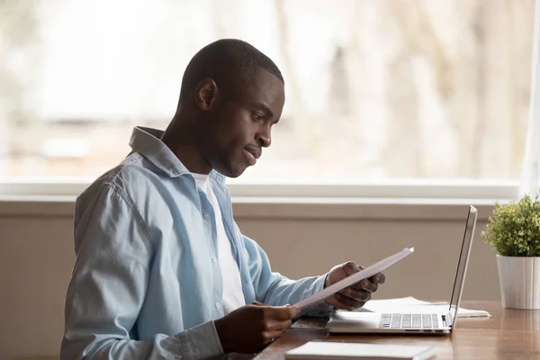 Focused biracial man reading paper letter working at laptop — Stock Photo, Image