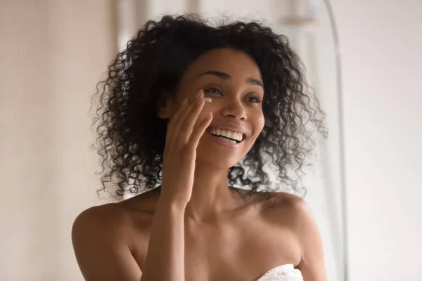 Sonriente mujer birracial aplicar bajo crema para los ojos mirando en el espejo —  Fotos de Stock