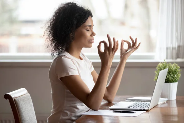 Calm biracial woman meditate near laptop at workplace