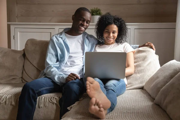 愉快的双种族夫妇放松在沙发上使用笔记本电脑 — 图库照片