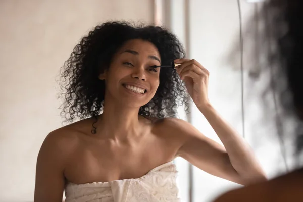 Gülümseyen biracial kadın banyoda maskara uygulayarak makyaj yapmak — Stok fotoğraf