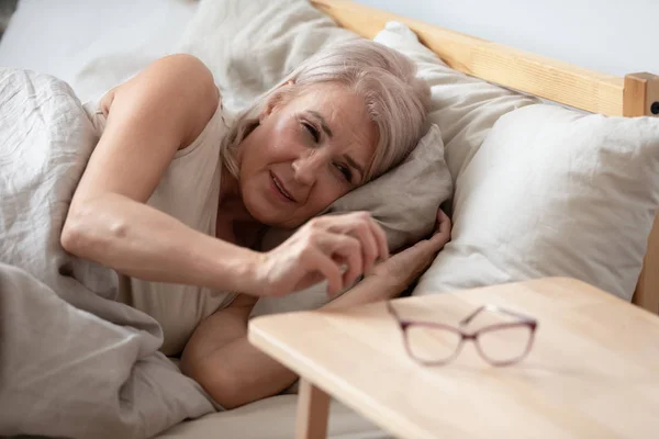 Ältere Frau nimmt morgens Gläser vom Nachttisch — Stockfoto