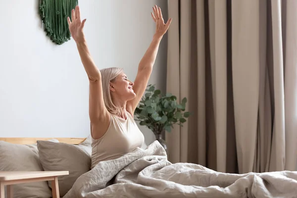 Mutlu yaşlı kadın yatakta oturan, uyandıktan sonra elleri germe — Stok fotoğraf