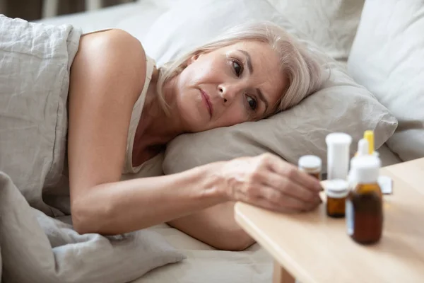 Unhealthy older woman taking pills from bedside table close up — Stock Photo, Image