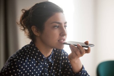 Ciddi Hintli genç iş kadını elinde telefonla dijital asistanıyla konuşuyor.