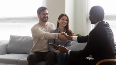 Gülümseyen genç evli çift Afrikalı Amerikalı emlakçıyla el sıkışıyor..