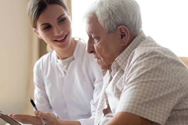 Uomo più anziano paziente e giovane infermiera parlando in casa — Foto Stock