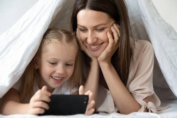 Happy mother and little daughter using phone, lying under blanket — Stock Photo, Image