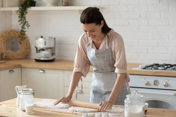 Mujer joven sonriente con delantal desplegando masa en la cocina —  Fotos de Stock