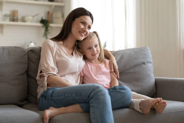 Щаслива молода мати обіймає маленьку доньку, сидячи на дивані разом — стокове фото