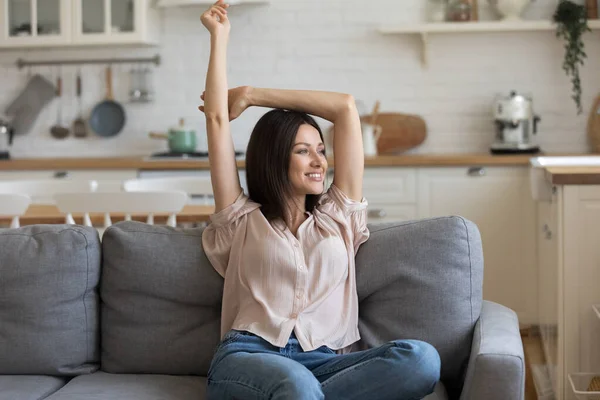 Smiling young woman relaxing on couch, stretching hands, looking aside — Stock Photo, Image