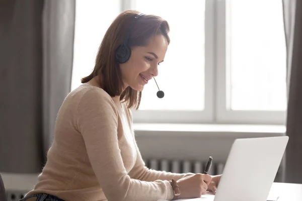 Uśmiechnięta bizneswoman w słuchawkach za pomocą laptopa, oglądając webinar, robiąc notatki — Zdjęcie stockowe