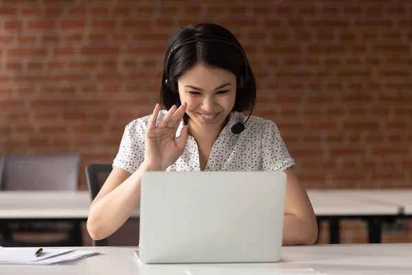 Smiling Asian businesswoman in headset waving hand, using laptop — Stock Photo, Image