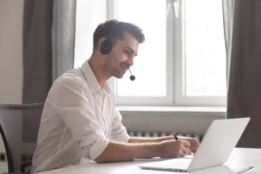 Kulaklıkla gülümseyen iş adamı dizüstü bilgisayar kullanıyor, Webinar izliyor, notlar alıyor.