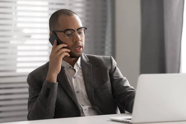 Serious African American businessman talking on phone, using laptop — Stock Photo, Image