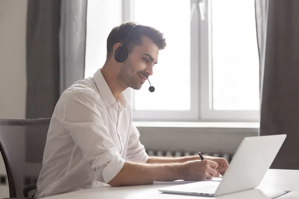 Smiling businessman in headset using laptop, watching webinar, making notes — Stock Photo, Image