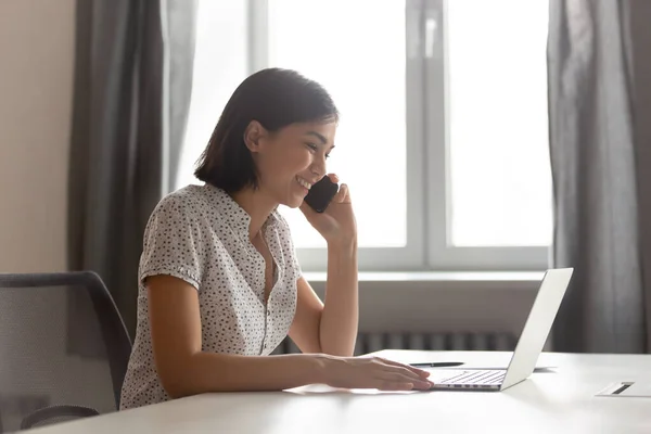 Smilende asiatisk forretningskvinne som snakker i telefonen og bruker laptop – stockfoto