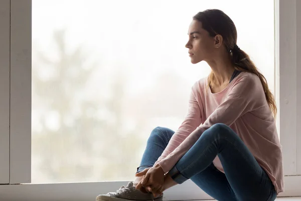 Sad pensive thoughtful girl sit on sill looking through window — Stock Photo, Image