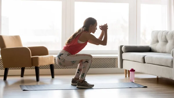 Mujer joven deportiva haciendo ejercicio en cuclillas por la mañana en la sala de estar — Foto de Stock