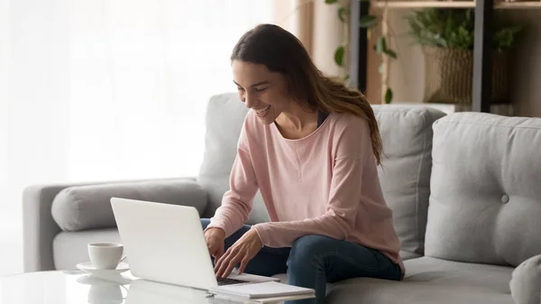 Kanepede oturup dizüstü bilgisayarla gülen mutlu genç bir kadın. — Stok fotoğraf
