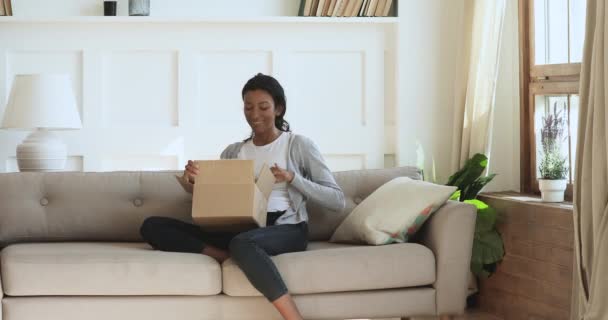 Nieuwsgierig gelukkig jong Afrikaans vrouw open perceel in woonkamer — Stockvideo