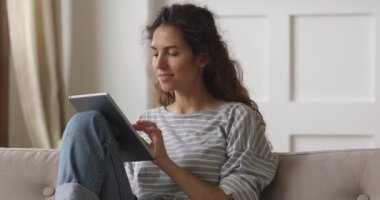 Mutlu genç kadın evinde kanepede tablet kullanarak dinleniyor.
