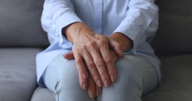 Yaşlı bir kadın kanepede elleri bağlı, yakın çekimde oturuyor.