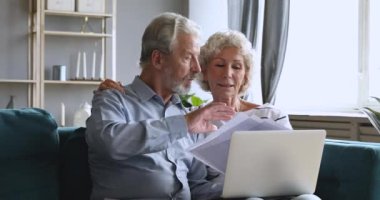 Ciddi, yaşlı bir çift ellerinde dizüstü bilgisayarlarıyla ev faturalarını kontrol ediyor.