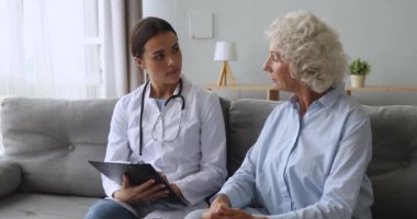 Genç kadın hemşire, not panosuna yazı yazıp kıdemli hastayla konuşuyor.