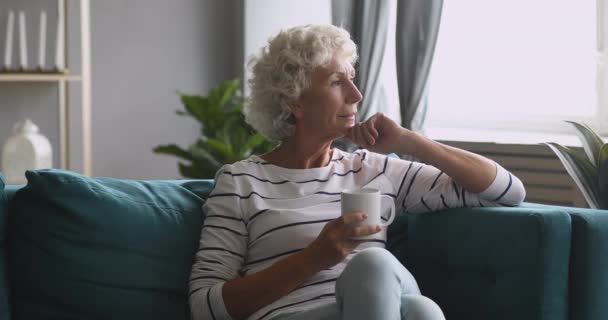 Allvarlig omtänksam gammal kvinna tittar bort dricka te på soffan — Stockvideo