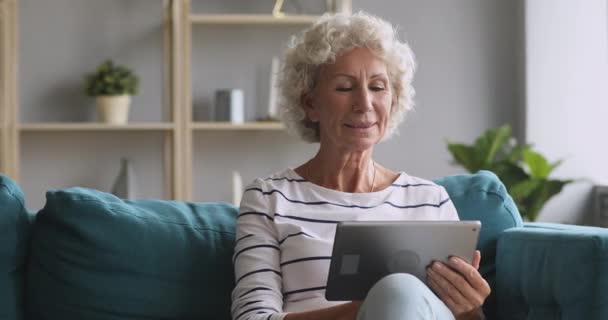 Улыбающаяся пожилая женщина с помощью цифрового планшета расслабляющий на диване — стоковое видео