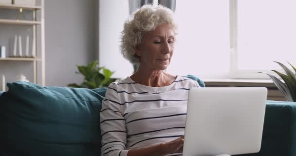 Doamnă în vârstă folosind tastarea laptopului pe computer așezat pe canapea — Videoclip de stoc