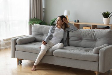 Positive dreamy young girl leaning on couch, holding phone.