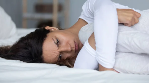 Депрессивная молодая смешанная расовая женщина лежит на кровати в спальне, обнимая колени . — стоковое фото