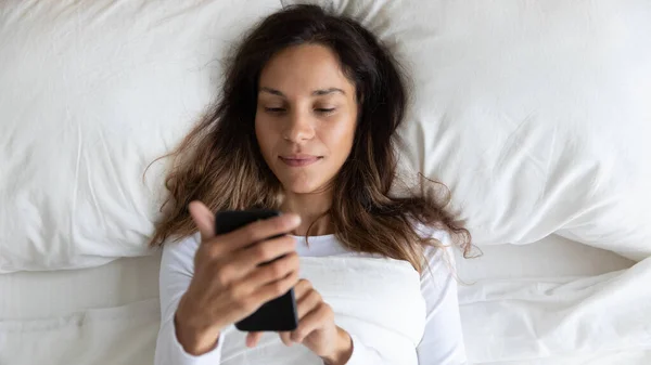 Pacifico joven mestiza chica despertando, usando el teléfono en la cama . — Foto de Stock