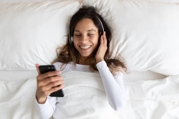 Boldog Y-generációs biracial nő fekszik az ágyban fülhallgatóval. — Stock Fotó