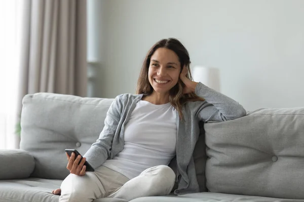 Glückliche Frau sitzt auf gemütlichem Sofa mit Smartphone in der Hand. — Stockfoto