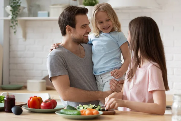 Usmívající se otec drží malou dceru, rodina připravuje večeři v kuchyni — Stock fotografie