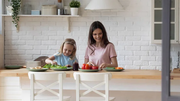 Gülümseyen anne ve kızı salata pişiriyor, mutfak masasında duruyorlar. — Stok fotoğraf