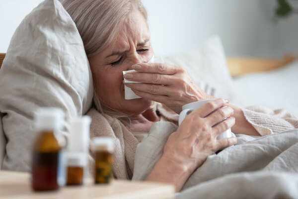 Ill middle aged woman sneezing blowing nose sit on bed