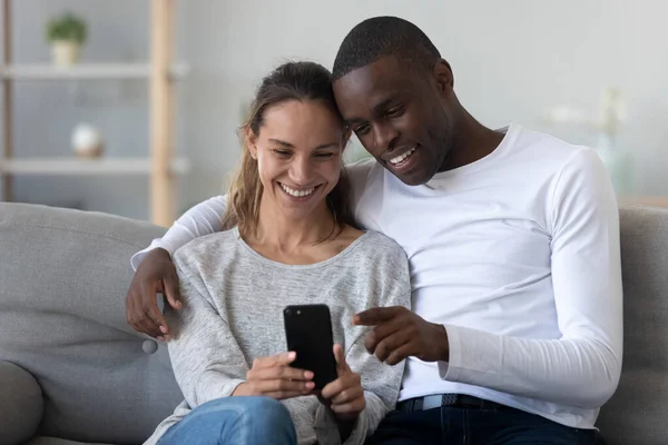 Feliz pareja internacional relajarse en el sofá utilizando el teléfono celular juntos —  Fotos de Stock