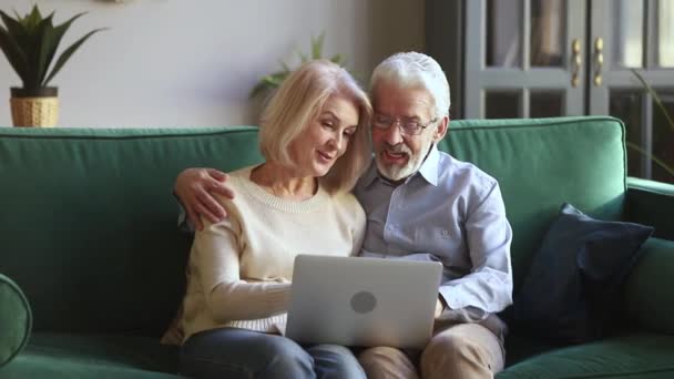 Sechzig Jahre positive fröhliche Eheleute, die mit Laptop auf der Couch sitzen — Stockvideo