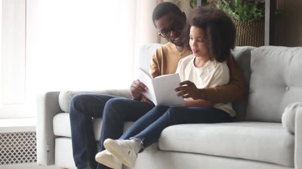 Afrikansk far läser bok för dotter sitter tillsammans på soffan — Stockvideo