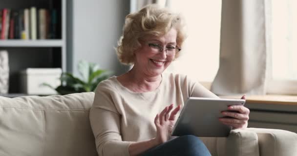 Uśmiechnięta starsza pani korzystająca z tabletu cyfrowego, relaksująca się na sofie. — Wideo stockowe