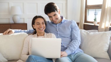 Gülümseyen genç çift evde birlikte dizüstü bilgisayar kullanarak rahatlıyor.