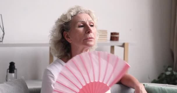 Беспокойная пожилая женщина использует бумажный вентилятор, чувствуя себя перегретой . — стоковое видео