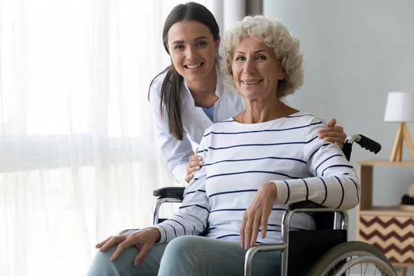 Caring nurse hug elderly disabled woman in wheelchair posing indoors — Stock Photo, Image